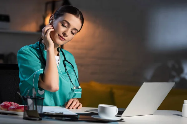 Attraente infermiera in uniforme seduta a tavola e ascoltare musica durante il turno di notte — Foto stock