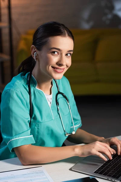 Attraktive und lächelnde Krankenschwester in Uniform mit Laptop und Musik während der Nachtschicht — Stockfoto