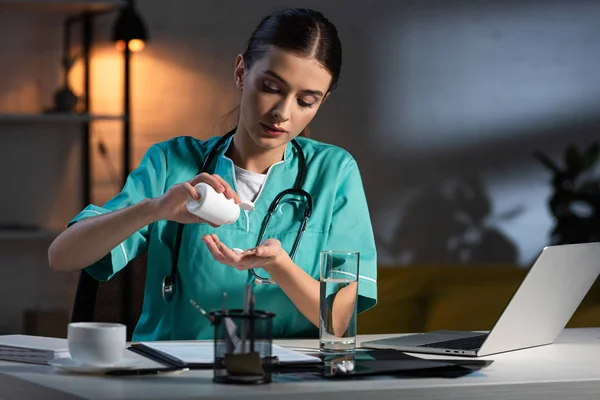Привлекательная медсестра в форме сидит за столом и принимает таблетки в ночную смену — стоковое фото