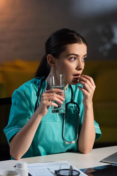 Enfermera atractiva en uniforme sentada en la mesa y tomando píldora durante el turno de noche - foto de stock