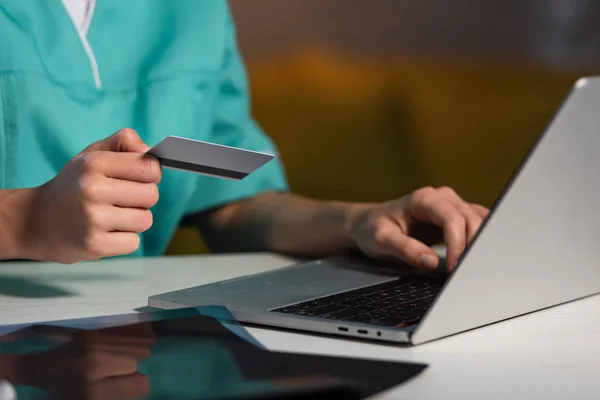 Cropped view of nurse in uniform holding credit card and using laptop during night shift — Stock Photo