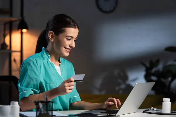 Усміхнена медсестра в уніформі медсестра в уніформі тримає кредитну картку і використовує ноутбук під час нічної зміни — стокове фото