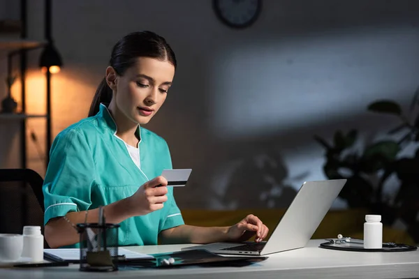 Enfermeira atraente em uniforme segurando cartão de crédito e usando laptop durante o turno da noite — Fotografia de Stock