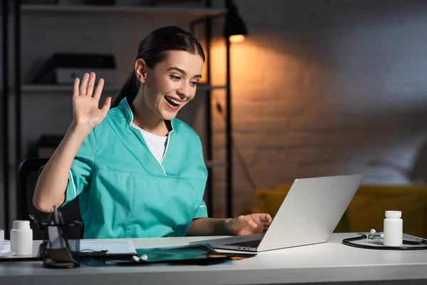 Attractive nurse in uniform sitting at table and having video chat during night shift — Stock Photo