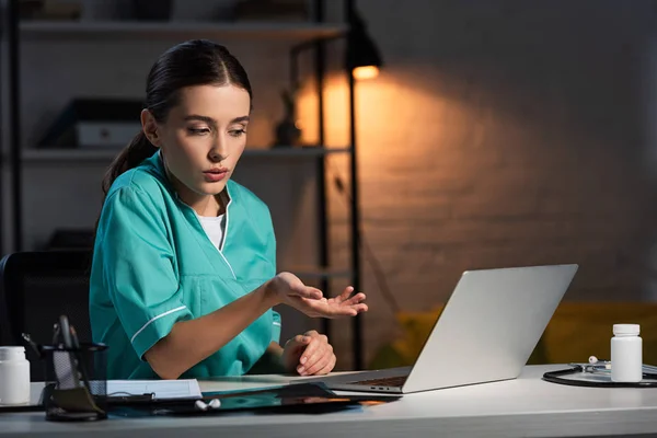 Enfermera atractiva en uniforme sentado en la mesa y tener video chat durante el turno de noche - foto de stock