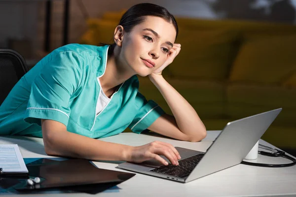 Attraente infermiera in uniforme seduta a tavola e utilizzando il computer portatile durante il turno di notte — Foto stock
