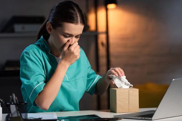 Attractive nurse in uniform sneezing and holding napkin during night shift — Stock Photo