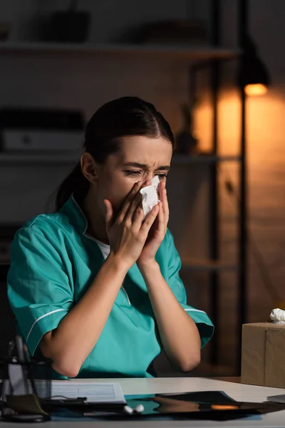 Attractive nurse in uniform sneezing and holding napkin during night shift — Stock Photo