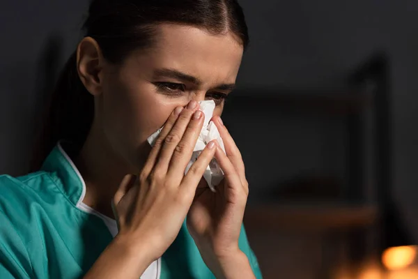 Enfermera atractiva en uniforme estornudando y sosteniendo la servilleta durante el turno de noche - foto de stock