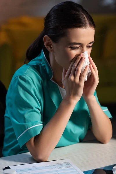 Attractive nurse in uniform sneezing and holding napkin during night shift — Stock Photo