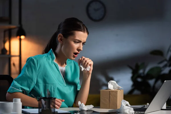 Attractive nurse in uniform sneezing and holding napkin during night shift — Stock Photo