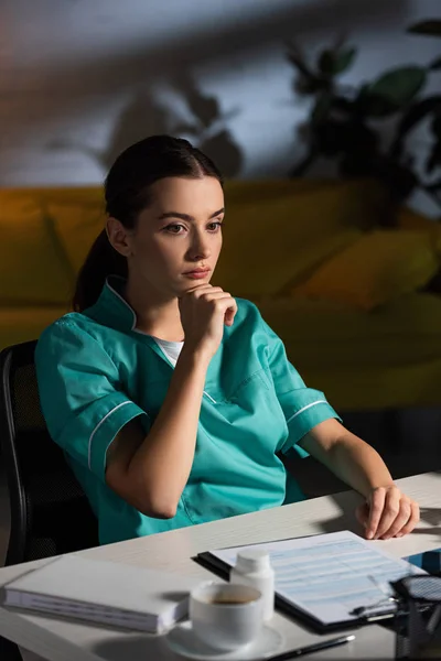 Attractive and pensive nurse in uniform sitting at table during night shift — Stock Photo