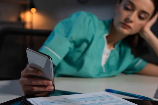 Messa a fuoco selettiva dell'infermiera in uniforme seduta a tavola e che utilizza lo smartphone durante il turno di notte — Foto stock