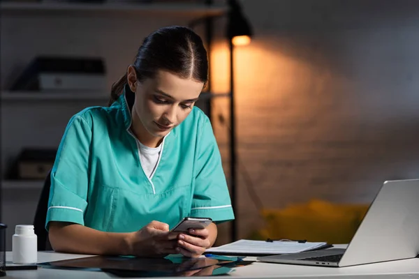 Infirmière attrayante en uniforme assis à table et utilisant un smartphone pendant le quart de nuit — Photo de stock