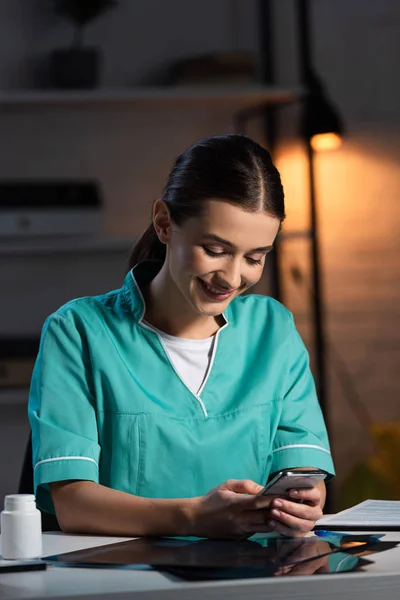 Infirmière attrayante en uniforme assis à table et utilisant un smartphone pendant le quart de nuit — Photo de stock