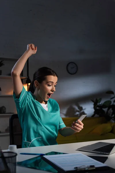Infirmière choquée en uniforme montrant geste oui et en utilisant smartphone pendant le quart de nuit — Photo de stock