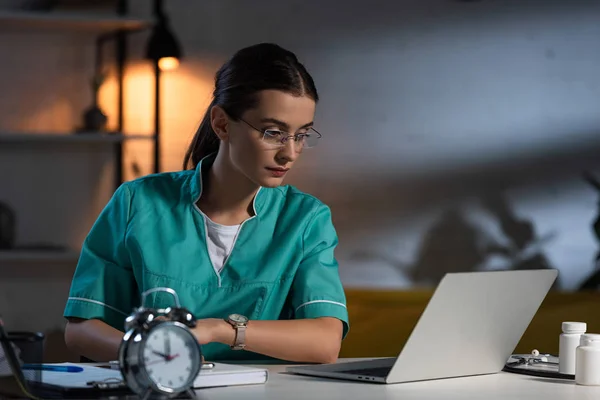 Enfermeira atraente em uniforme e óculos sentados à mesa e olhando para laptop durante o turno da noite — Fotografia de Stock