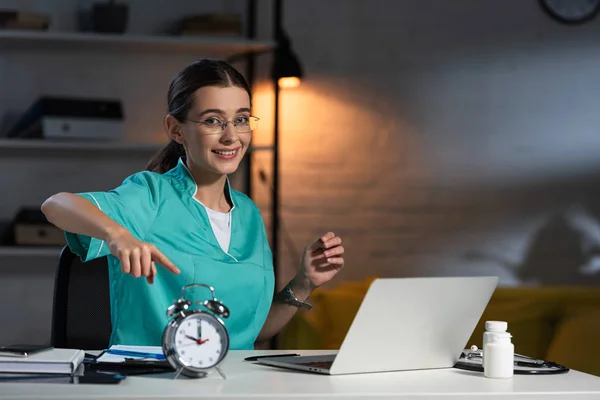 Attractive nurse in uniform and glasses sitting at table and pointing with finger at alarm watch during night shift — Stock Photo