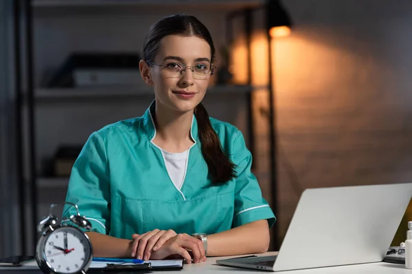 Attraktive Krankenschwester in Uniform und Brille, die am Tisch sitzt und während der Nachtschicht in die Kamera schaut — Stockfoto