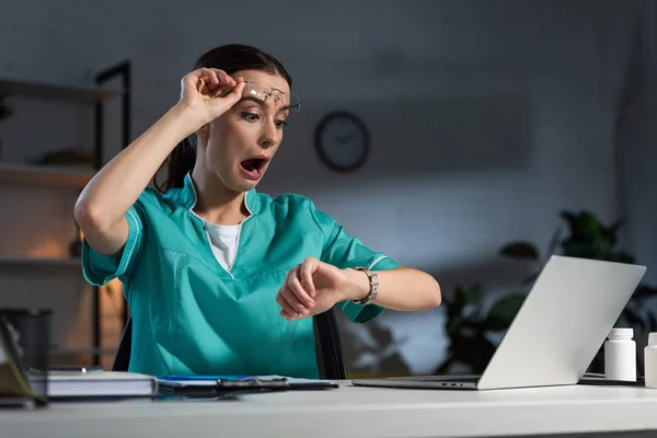 Schockierte Krankenschwester in Uniform und Brille sitzt am Tisch und schaut während der Nachtschicht auf die Armbanduhr — Stockfoto