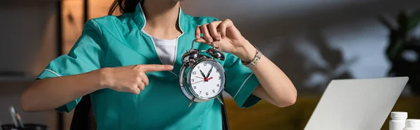 Panoramic shot of nurse in uniform pointing with finger at alarm watch during night shift — Stock Photo