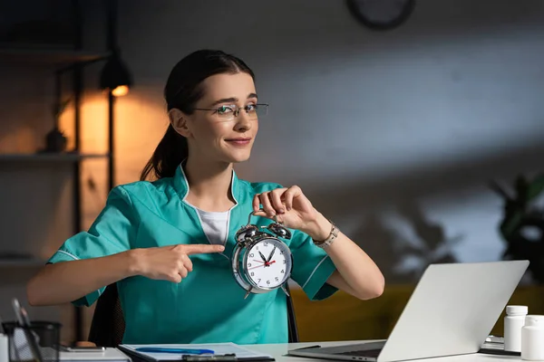 Attraente infermiera in uniforme e occhiali seduti a tavola e che punta con il dito all'orologio di allarme durante il turno di notte — Foto stock