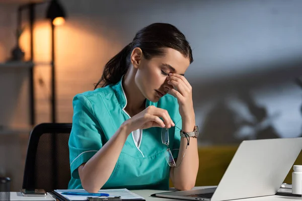 Attractive nurse in uniform holding glasses and sitting at table during night shift — Stock Photo