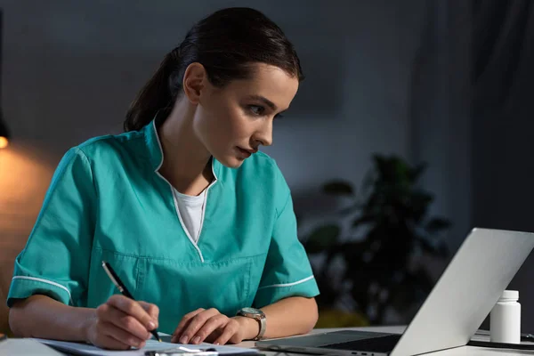 Attraktive Krankenschwester in Uniform, die während der Nachtschicht am Tisch sitzt und schreibt — Stockfoto