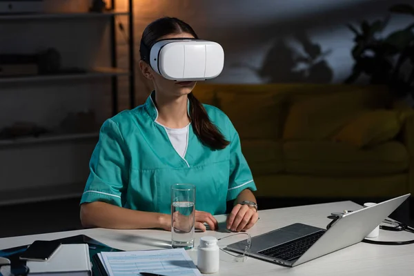 Infirmière en uniforme avec casque de réalité virtuelle assise à table pendant le quart de nuit — Photo de stock