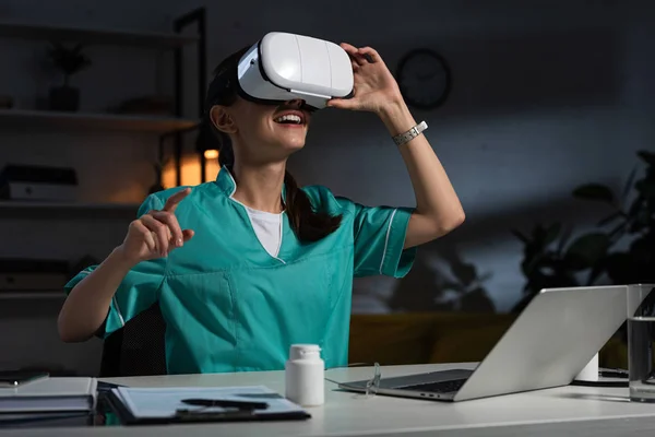 Nurse in uniform with virtual reality headset pointing with finger during night shift — Stock Photo