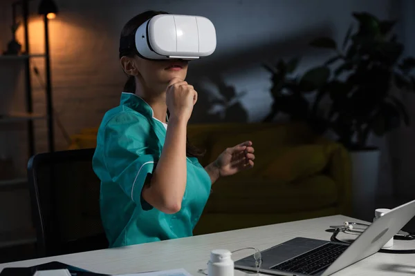 Enfermera pensativa en uniforme con auriculares de realidad virtual sentado en la mesa durante el turno de noche - foto de stock