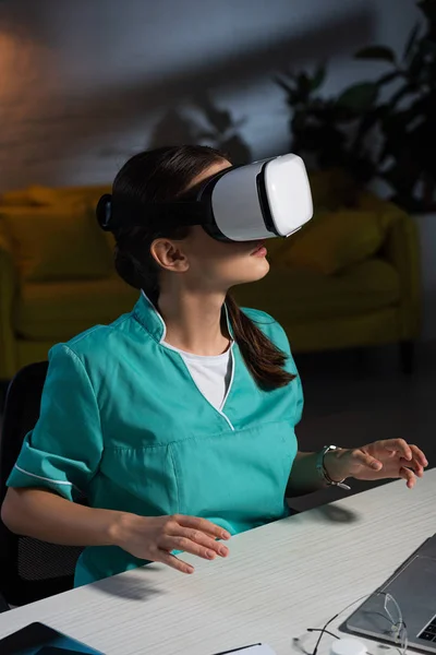 Infirmière en uniforme avec casque de réalité virtuelle assise à table pendant le quart de nuit — Photo de stock