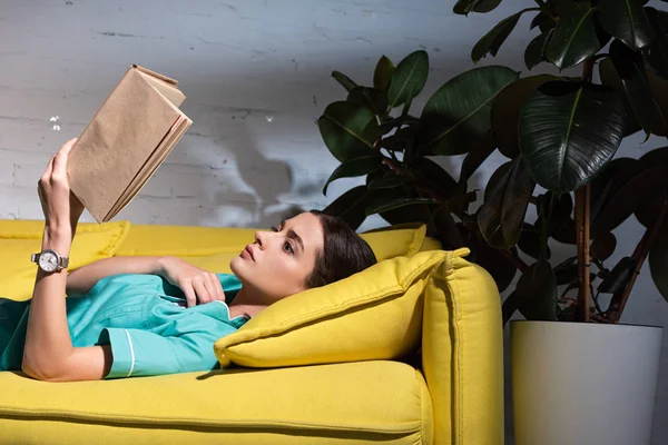 Infirmière attrayante en uniforme couché sur le canapé et le livre de lecture pendant le quart de nuit — Photo de stock