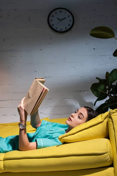 Infirmière attrayante en uniforme couché sur le canapé et le livre de lecture pendant le quart de nuit — Photo de stock