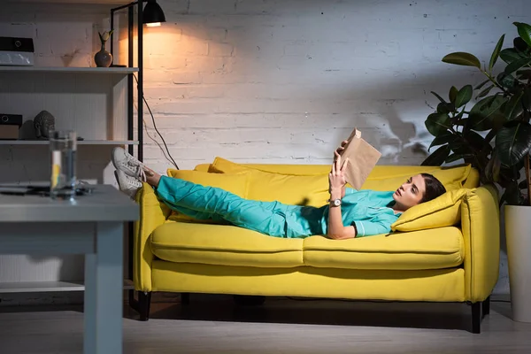 Infirmière attrayante en uniforme couché sur le canapé et le livre de lecture pendant le quart de nuit — Photo de stock