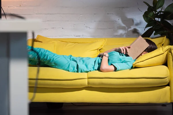 Foyer sélectif de l'infirmière en uniforme couché sur le canapé avec livre sur le visage pendant le quart de nuit — Photo de stock