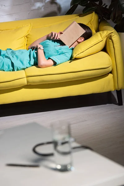 Selective focus of nurse in uniform lying on sofa with book on face during night shift — Stock Photo