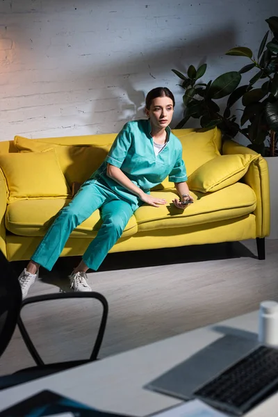 Infirmière en uniforme assise sur le canapé et regardant ailleurs pendant le quart de nuit — Photo de stock