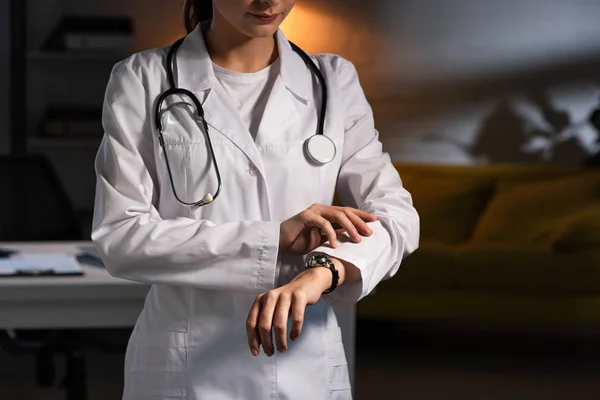 Vue recadrée du médecin en manteau blanc avec stéthoscope regardant la montre-bracelet pendant le quart de nuit — Photo de stock