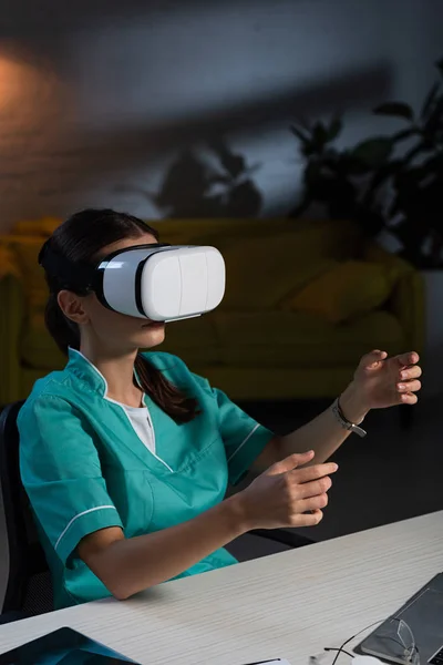 Nurse in uniform sitting at table with virtual reality headset during night shift — Stock Photo