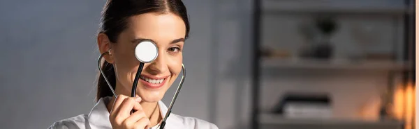 Panoramic shot of doctor in white coat holding stethoscope during night shift — Stock Photo