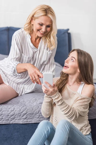 Mulher madura feliz sentada na cama e apontando com o dedo para o smartphone em mãos de filha alegre jovem — Fotografia de Stock