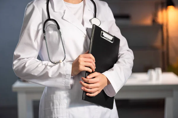 Vista recortada del médico de bata blanca con estetoscopio sujetando el portapapeles durante el turno de noche - foto de stock