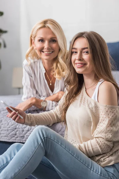 Cheerful girl holding smartphone and mother lying on bed and smiling at camera — Stock Photo