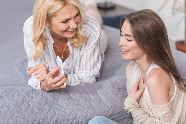Lachendes Mädchen blickt auf Smartphone neben glücklicher Mutter, die im Bett liegt — Stockfoto