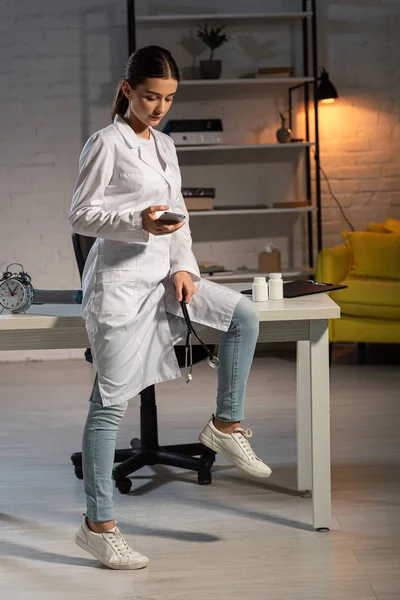 Attraktiver Arzt in weißem Mantel, Smartphone in der Hand und während der Nachtschicht auf dem Tisch sitzend — Stockfoto