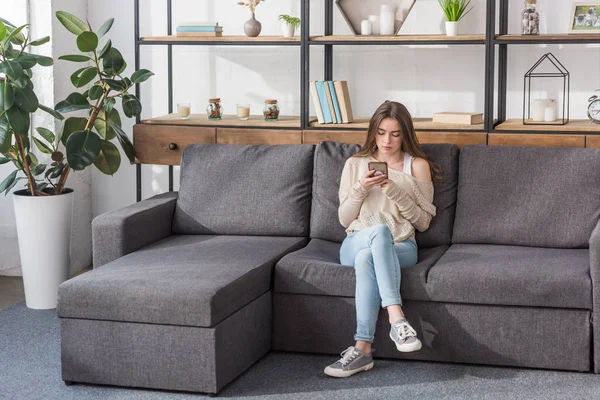 Pretty girl using smartphone while sitting on sofa in living room — Stock Photo