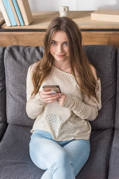 Jolie, fille souriante regardant la caméra tout en étant assis sur le canapé et en utilisant un smartphone — Photo de stock