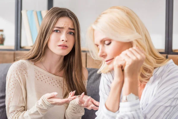 Offended girl looking at camera and showing question gesture while sitting near upset mother — Stock Photo