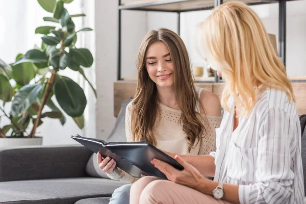 Pretty girl smiling while sitting on sofa with mother and looking at photo album — Stock Photo